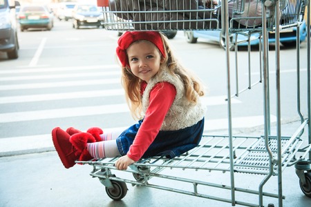 A little sweet girl is sitting in a supermarket shopping cart ootdoors.の写真素材