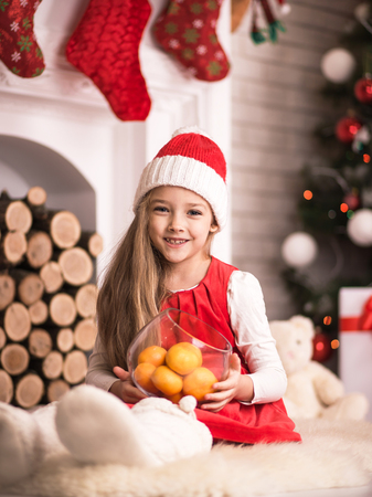 Lovely girl in santa cap with tangerines in hand, sitting on floor in red dress, studio shot, toning in vintage style.の写真素材