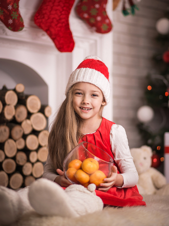 Lovely girl in santa cap with tangerines in hand, sitting on floor in red dress, studio shot.の写真素材