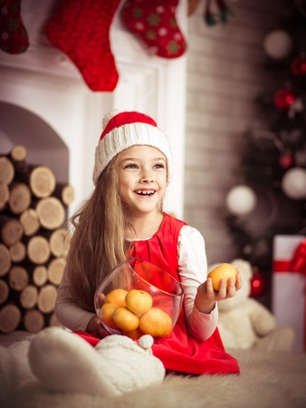 Lovely girl in santa cap with tangerines in hand, sitting on floor in red dress, studio shot.の写真素材
