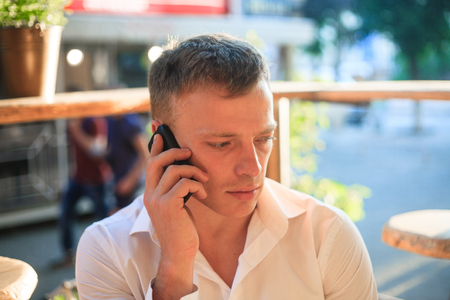 Portrait of a young man with a phone in hands in a street cafe.の写真素材