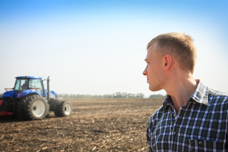 Young man in a field and a tractor on a background. Concept of agriculture and field works.の写真素材