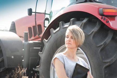Pretty young woman with tablet standing in field with tractor in backgroundの写真素材