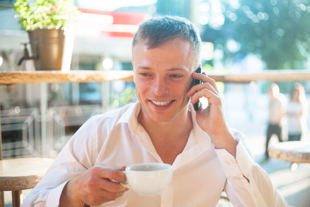 Young man drinking coffee in cafe and using phone. Business people outdoors portrait.の写真素材