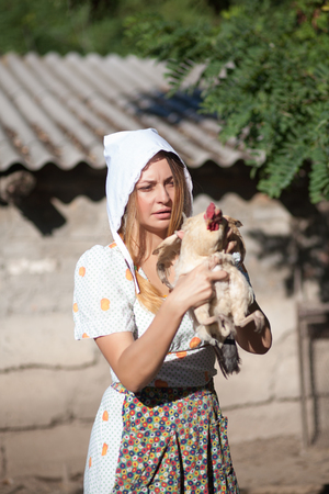 A woman in a village dressed in country style is holding a hen. Shooting in retro style.の写真素材