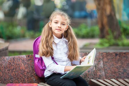 Little school girl with a backpack and book walking outdoors after lessons, read book or study, education and learning concept.の写真素材