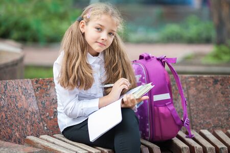 Little school girl with a backpack and book walking outdoors after lessons, read book or study, education and learning concept.の写真素材