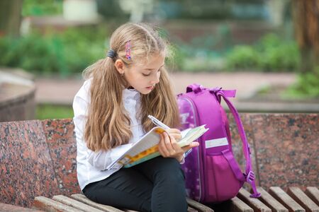 Little school girl with a backpack and book walking outdoors after lessons, read book or study, education and learning concept.の写真素材