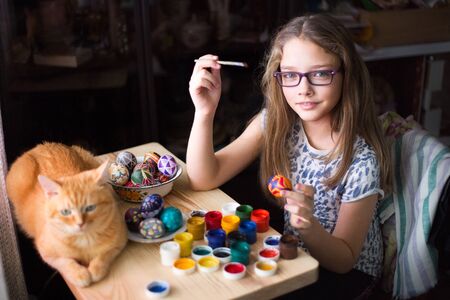 Teen girl paints Easter eggs, her ginger cat lies on the table. Home interior, easter holiday.の写真素材