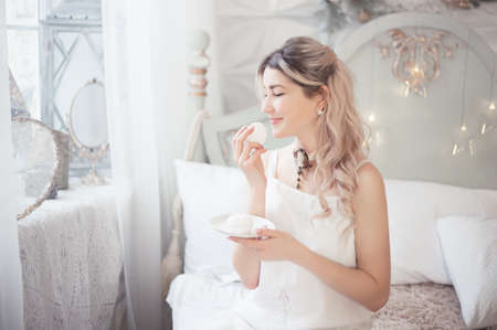 Cute young woman eating sweets in the morning in the bedroom. Gentle winter morning. Studio photo.の写真素材