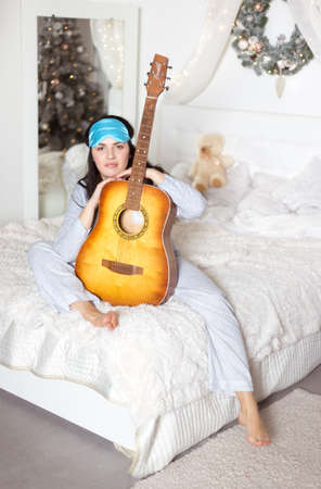 Cute beautiful woman in bed in pajamas with a guitar in her hands. Winter holidays, christmas, studio photo.の写真素材