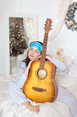 Cute beautiful woman in bed in pajamas with a guitar in her hands. Winter holidays, christmas, studio photo.の写真素材