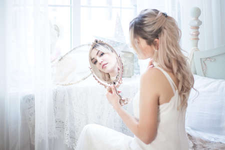 Young beautiful woman looks in the mirror in her bedroom. Morning of the bride.の写真素材