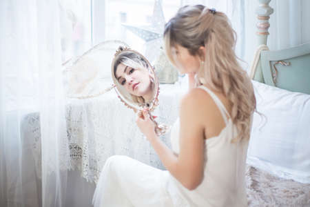 Young beautiful woman looks in the mirror in her bedroom. Morning of the bride.の写真素材