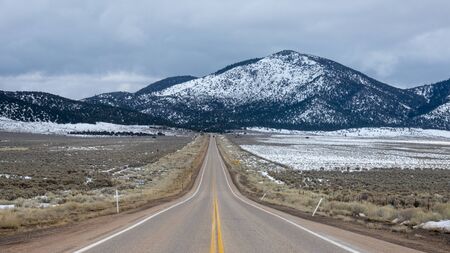 Empty highway along dull frozen field near against mountain range covered with snowの写真素材