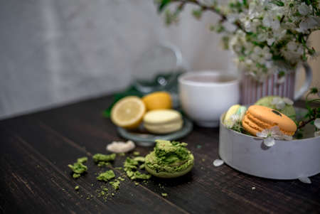Crushed pastel, orange and green macaroons on a wood background with whole one in white plate with jug with flowers on the backの写真素材