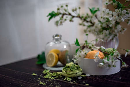 Crushed pastel, orange and green macaroons on a wood background with whole one in white plate with jug with flowers on the backの写真素材