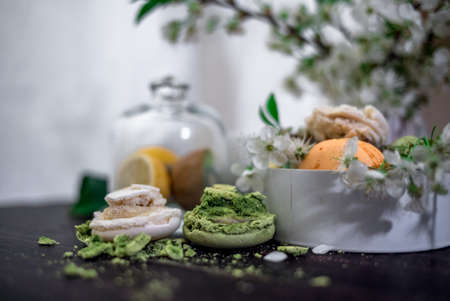 Crushed pastel, orange and green macaroons on a wood background with whole one in white plate with jug with flowers on the backの写真素材