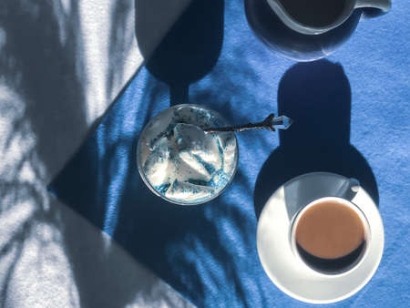 Coffee in a white cup with a spoon,  Ice cream in a glass and small jug of milk on a blue and white background with dry wild grass shadowsの写真素材