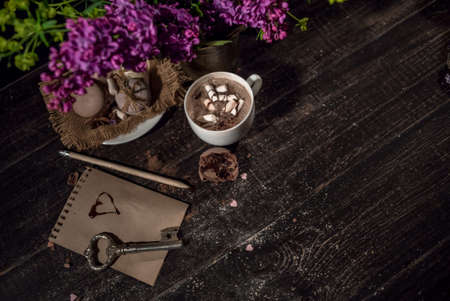 Hot chocolate (cocoa) in a glass mug with two tubes for drinking.There are some macarons on a linen napkin on the wood table with some lilacの写真素材