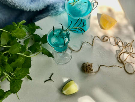 Blue cold drink in a glass with ice cubes on white desk with blue background. Also used mint leaves and rosemary, rope, spoon  with natual sunlight from the open windowの写真素材