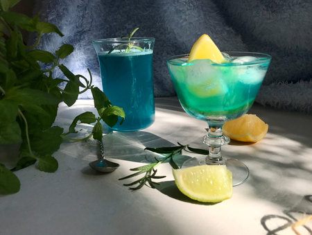 Blue cold drink in a glass with ice cubes on white desk with blue background. Also used mint leaves and rosemary, rope, spoon  with natual sunlight from the open windowの写真素材