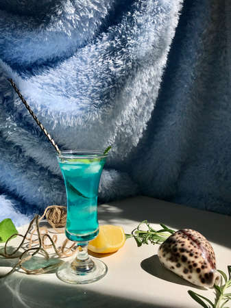 Blue cold drink in a glass with ice cubes on white desk with blue background. Also used mint leaves and rosemary, rope, spoon  with natual sunlight from the open windowの写真素材