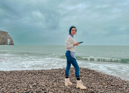 Young woman standing on the beach with mobile phone in her hand. Young woman with mobile phone in...