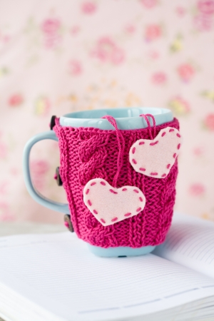 Blue cup in pink sweater with felt hearts standing on an open notebookの写真素材