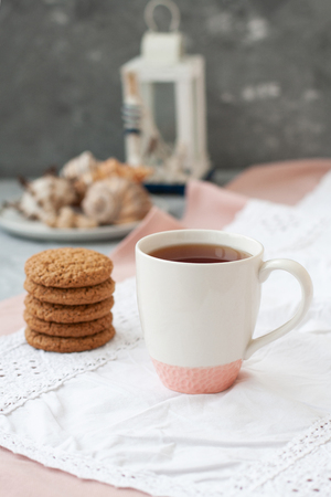 A tasty snack: a cup of black tea, a stack of oatmeal cookies; a decorative lantern and seashells on the gray background.の写真素材