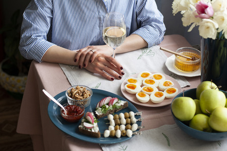 A gourmet lunch for two: rye bread sandwiches with cream cheese and radish, spicy sauce, a bowl of fruits; boiled eggs, mozzarella and olives canapes; bowls with honey and walnuts, a glass of wine; a vase with flowers on a pink tablecloth.の写真素材
