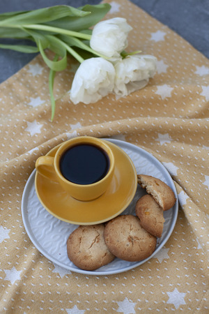 A tasty snack: a cup of black coffee, a plate of cookies; a bouquet of white roses on a golden tablecloth on a gray background.の写真素材