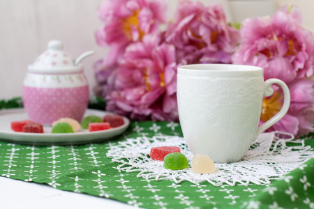 A tasty break: a cup of tea with jelly beans of various tastes, a sugar bowl on green decorative napkin; a bouquet of pink flowers on a white background.の写真素材