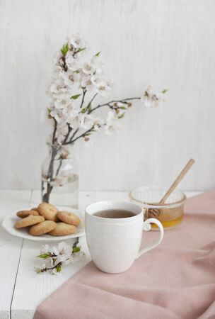A tasty break: a cup of black tea on a pink napkin, a bowl of honey, a plate of homemade cookies; a glass vase of apricot bloom on the white background.の写真素材