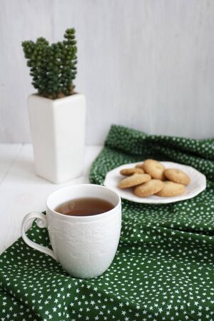 A tasty break: a cup of black tea on a green napkin, a plate of homemade cookies; a decorative plant in a vase on the white background.の写真素材