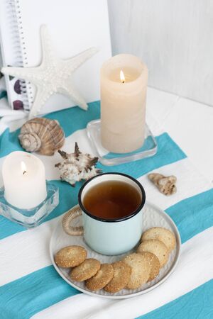 A study break: a hot mug of black tea with homemade cookies on a striped tablecloth; wax candles, decorative starfish, seashell, pile of notebooks on a white background.の写真素材