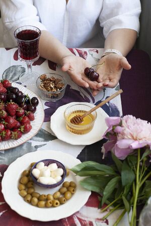 A gourmet dinner for two: grilled camambert with armula and walnuts on top, a glass of red wine, a plate of cherries and strawberries, a plate olives with mozzarella balls, bowls of honey and walnuts; a vase with flowers on a decorative tablecloth.の写真素材