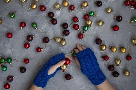 Hands of young beautiful woman in blue mittens with a pattern. She holds a green ball in one hand and a red and yellow ball in the other. composition on a gray concrete background. Around there are red, green, cherry and gold ornaments on the Christmas tree.の写真素材