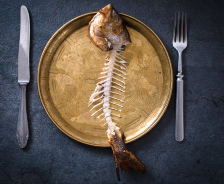 Fish skeleton on plate with fork and knife on dark background, top viewの写真素材