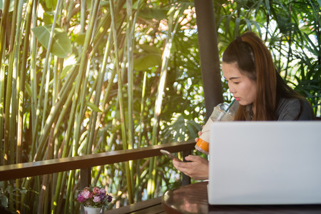 Woman hold mobile phone and drinking iced tea in coffee shop, laptop on wooden table of brownの写真素材