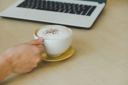Closeup image of woman hand holding a cup of hot coffee on wooden table, working with computer laptop, freelance and work conceptの写真素材