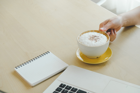 Closeup image of woman hand holding a cup of hot coffee on wooden table, working with computer laptop, freelance and work conceptの写真素材