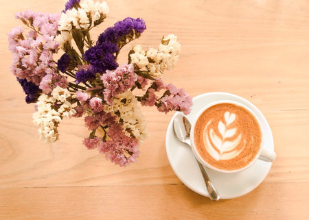 A cup of latte or cappuccino with statice or limonium flower on wooden spruce desk with bright retro...