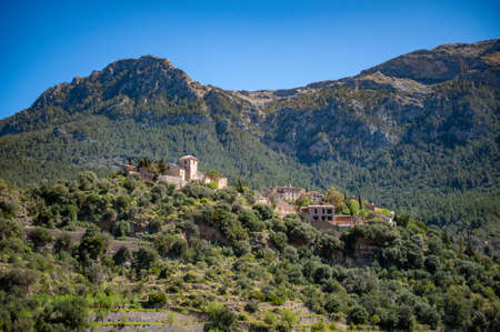 Spanish village on a green forested mountain. Behind it stretches a higher mountain range. Blue, cloudless sky.の写真素材