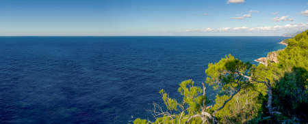 We take a break and look down from a mountain at the wide, deep blue sea. Rocks with vegetation can be seen at the edge. A light sea breeze blows around our noses.の写真素材