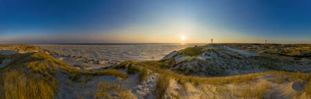 Panoramic shot of a wide landscape covered with dune grass overlooking the sea. Evening sky at sunset. A lighthouse can be seen small.の写真素材
