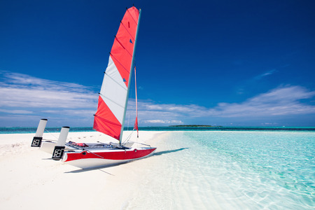 Sailing boat with red sail on a beach of deserted tropical island with shallow blue waterの写真素材
