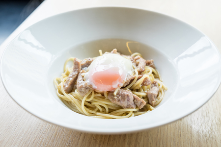 Hot Spaghetti and fried sliced pork with garlic top up with onsen egg on white plate and wooden table, Thai and  Italian fusion food styleの写真素材