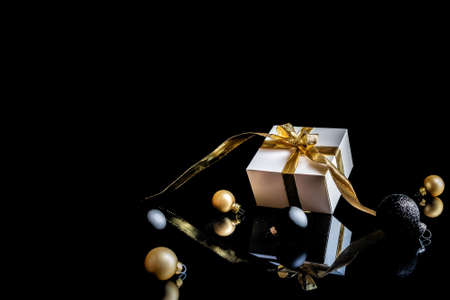 Gift ribbon gold. White gift box with golden ribbon, New Year balls and winter tree in Christmas composition on black background for greeting card. Winter festive composition with copy spaceの写真素材