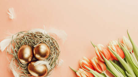 Egg color. Happy Easter decoration: golden color eggs in basket with spring tulips, white feathers on pastel pink background. Foil minimalist egg design, modern top view templateの写真素材